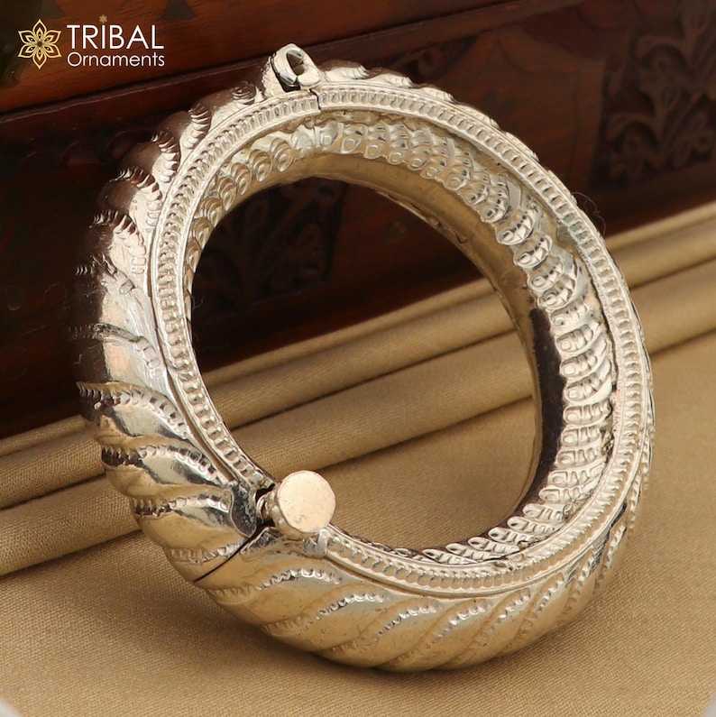 May include: A silver bangle bracelet with intricate, detailed designs. The bracelet is hinged and opens to fit the wrist.