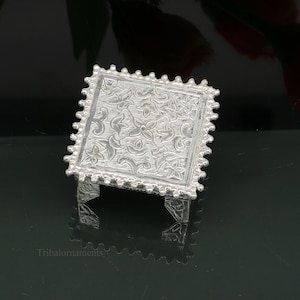 Puede incluir: Un soporte decorativo cuadrado de plata con un intrincado diseño floral y un borde de cuentas. El soporte tiene cuatro patas y está ligeramente elevado de la superficie.