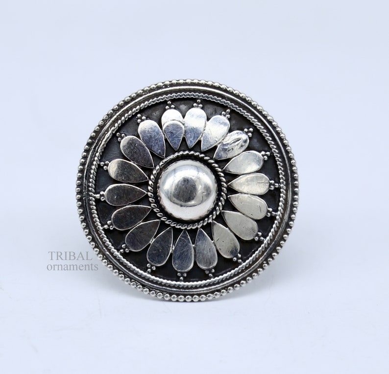 May include: A silver ring with a circular design featuring a central dome surrounded by a ring of petals. The ring has a textured, beaded edge.