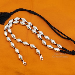 May include: A silver necklace with a black cord and silver beads. The necklace has a tassel on one end and two silver clasps on the other. The necklace is on a orange background. TRIBAL Ornaments is written on the background.
