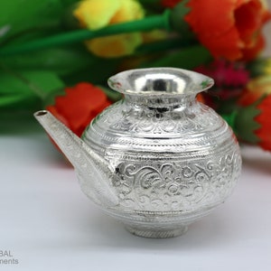 May include: A silver, intricately engraved, traditional Indian water pot with a spout. The pot is round with a wide base and a narrow neck. It is decorated with floral patterns.