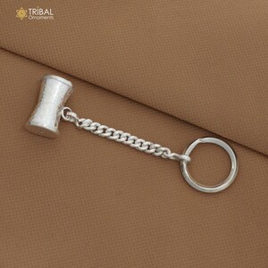 May include: A silver-colored keychain featuring a small, textured hammer-shaped charm connected to a chain and a round key ring. The keychain has the text "TRIBAL Ornaments" in the upper left corner.