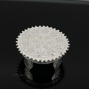 Puede incluir: Un soporte decorativo redondo plateado con un patrón floral en la superficie superior. El soporte tiene tres patas cortas y un borde con cuentas. El fondo es negro.