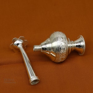 May include: A silver, ornate, decorative bottle with a floral design and a separate nozzle. The bottle is lying on a brown surface.