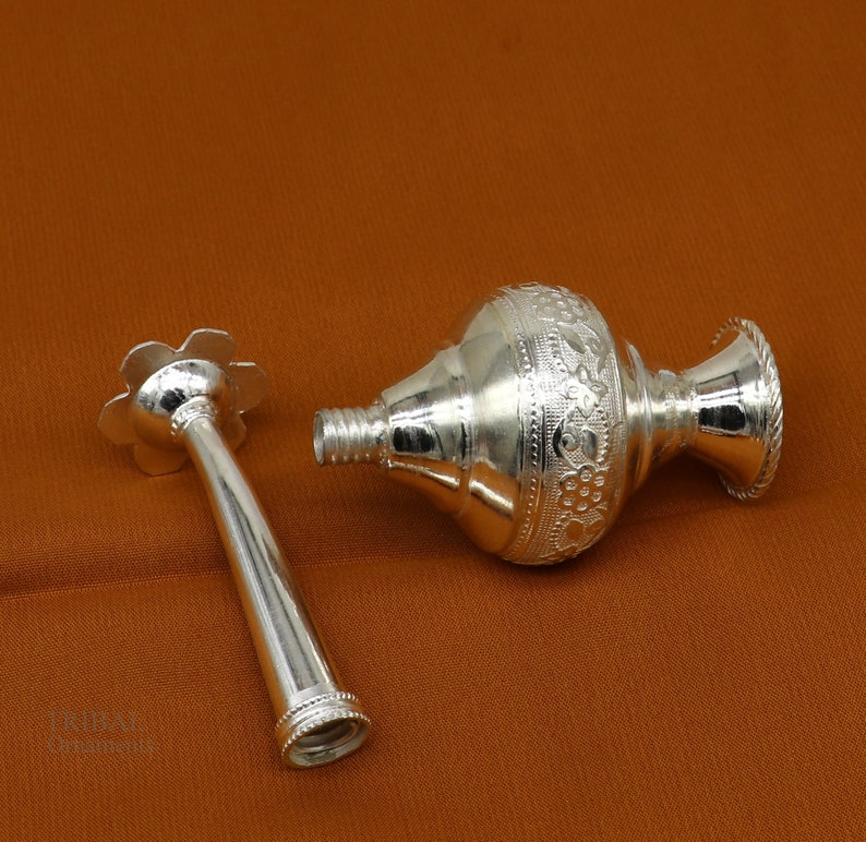 May include: A silver, ornate, decorative bottle with a floral design and a separate nozzle. The bottle is lying on a brown surface.