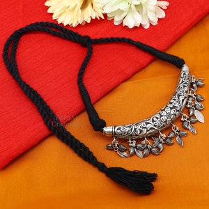 May include: A silver-colored necklace with a floral design and dangling leaf charms. The necklace has a black rope cord and tassel. The background is a combination of red and orange fabric.
