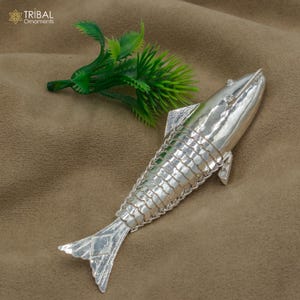 May include: A silver fish ornament with a detailed, textured surface. The fish is lying on a brown fabric background.
