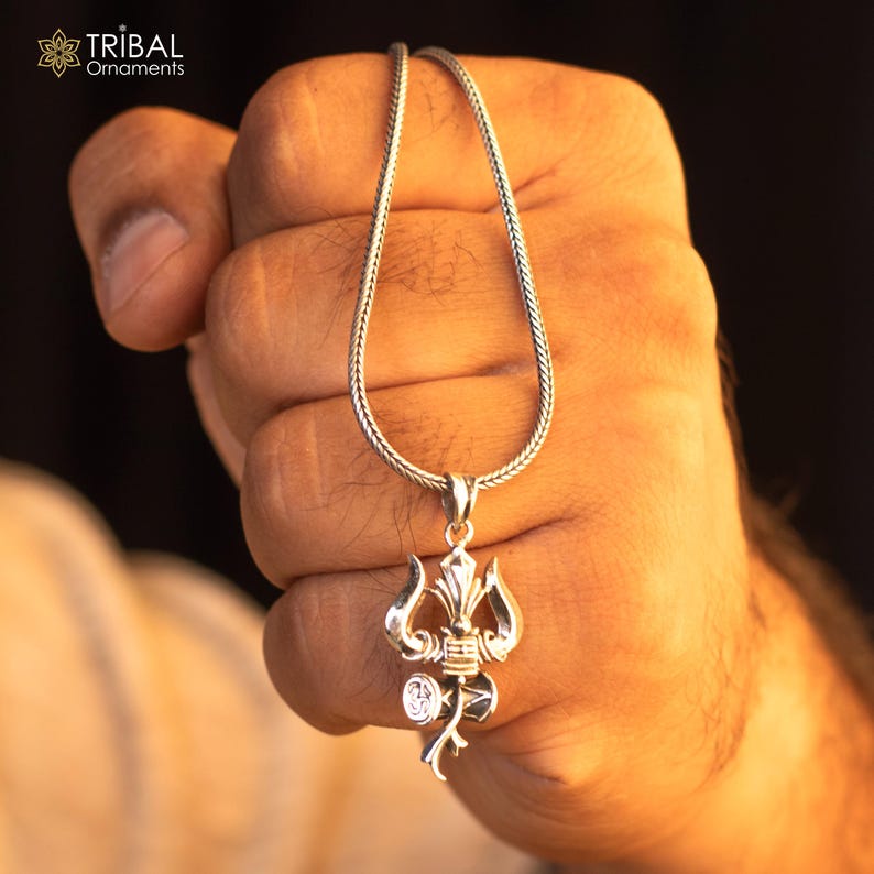 May include: A silver chain necklace with a silver pendant featuring a trident design. The pendant is detailed with a small circle and a stylized design.