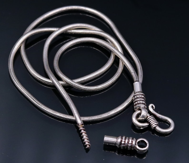 May include: A silver snake chain necklace with a hook clasp and a small, round connector. The chain is coiled on a black surface.