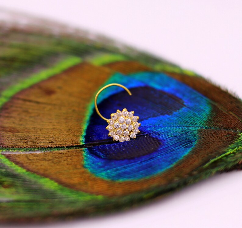 Puede incluir: Un aro de nariz de oro con un c&uacute;mulo redondo de piedras de circonita c&uacute;bica transparentes. El aro est&aacute; apoyado sobre una pluma de pavo real.