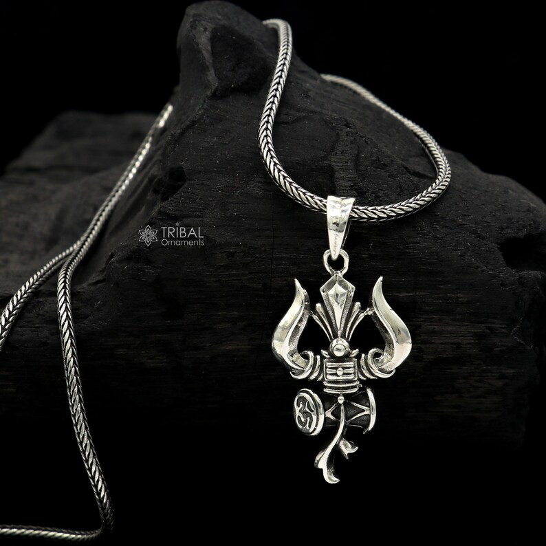 May include: A silver necklace with a detailed trident pendant. The pendant features a three-pronged spear with a circular design at the base. The necklace is made of a silver chain.