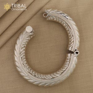 May include: Silver bangle bracelet with an intricate, ornate design. The bracelet is open and has a hinged clasp.