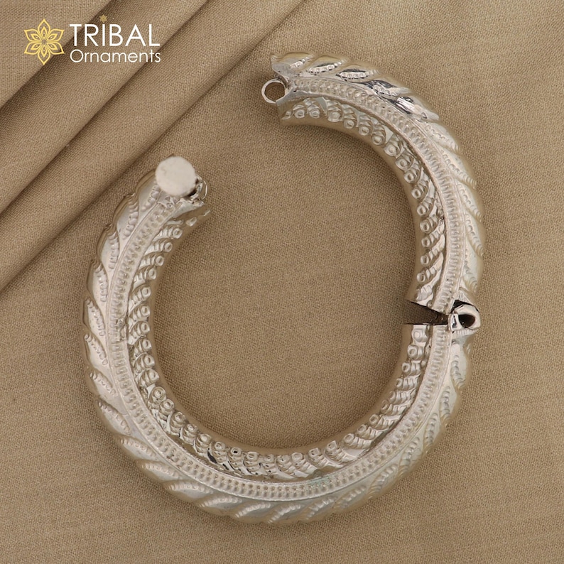 May include: Silver bangle bracelet with an intricate, ornate design. The bracelet is open and has a hinged clasp.