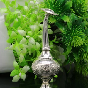 May include: A silver decorative bottle with an ornate design and a long, curved spout. The bottle is sitting on a black surface with green plants in the background.