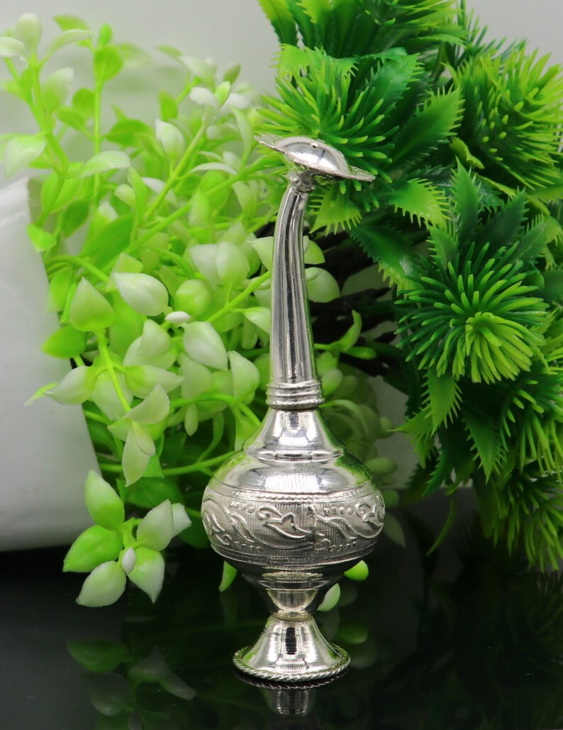 May include: A silver decorative bottle with an ornate design and a long, curved spout. The bottle is sitting on a black surface with green plants in the background.