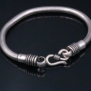 K&ouml;nnte beinhalten: Ein silbernes Schlangenkettenarmband mit einem Hakenverschluss. Das Armband besteht aus strukturiertem Metall und hat eine polierte Oberfl&auml;che.