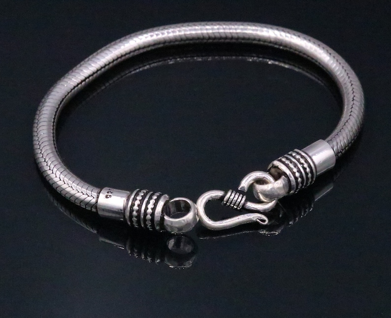K&ouml;nnte beinhalten: Ein silbernes Schlangenkettenarmband mit einem Hakenverschluss. Das Armband besteht aus strukturiertem Metall und hat eine polierte Oberfl&auml;che.