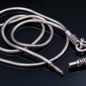 May include: A silver snake chain necklace with a clasp and a connector. The chain is coiled and laid out on a black surface.