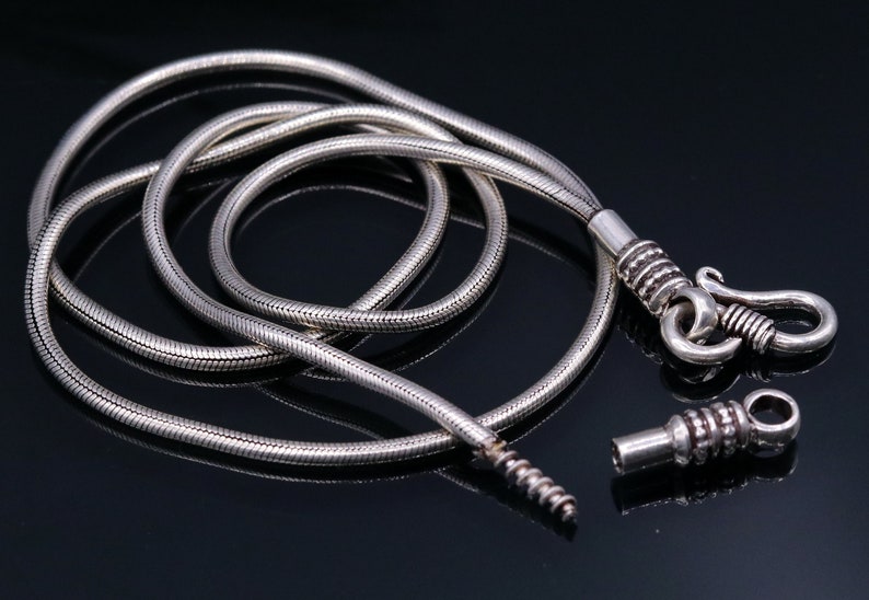 May include: A silver snake chain necklace with a clasp and a connector. The chain is coiled and laid out on a black surface.
