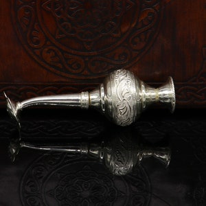 May include: A silver, ornate, decorative bottle with a long, thin spout. The bottle has an intricate floral design and is resting on a dark brown surface.