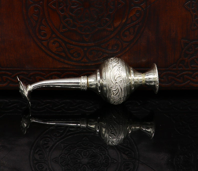 May include: A silver, ornate, decorative bottle with a long, thin spout. The bottle has an intricate floral design and is resting on a dark brown surface.