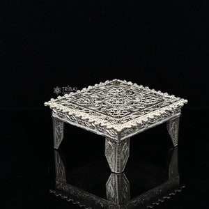 Puede incluir: Un pedestal cuadrado de plata con intrincados diseños florales y un borde de cuentas. El pedestal tiene cuatro patas y está hecho de metal.