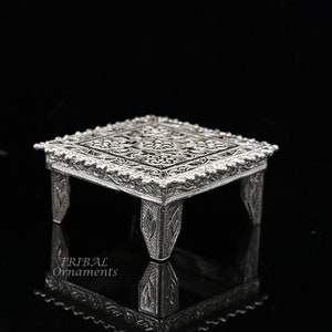 Puede incluir: Un pedestal cuadrado de plata, con un diseño intrincado, con cuatro patas. El pedestal tiene un patrón floral decorativo.
