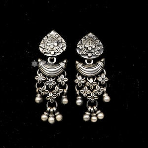 May include: A pair of silver earrings with a floral design. Each earring features a teardrop-shaped top with a detailed floral pattern, a curved middle section, and a dangling cluster of flowers and small beads. The earrings are set against a black background.