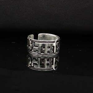 May include: Silver ring with a decorative design featuring a repeating pattern of letters. The ring is wide and has a slightly rounded shape.
