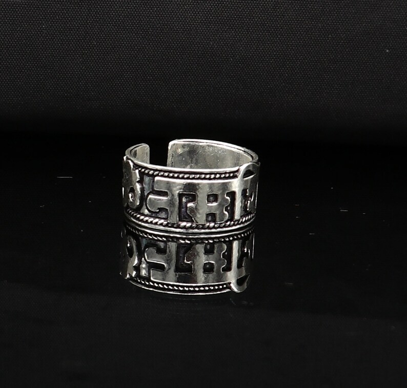 May include: Silver ring with a decorative design featuring a repeating pattern of letters. The ring is wide and has a slightly rounded shape.
