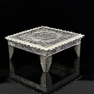 Puede incluir: Un pedestal cuadrado de plata con intrincados diseños florales y un centro elevado. El pedestal tiene cuatro patas y está hecho de metal.