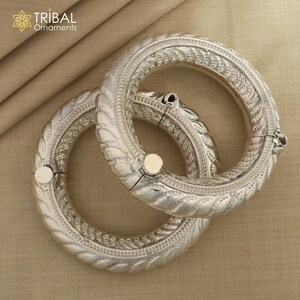May include: Two silver bangles with intricate designs. The bangles are made of metal and have a shiny finish. The bangles are stacked on top of each other.