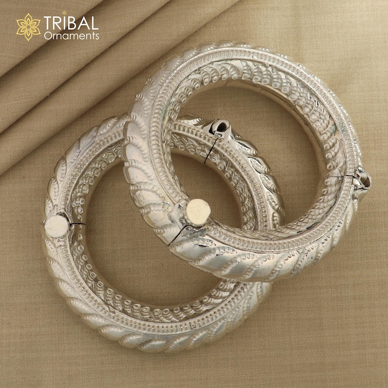 May include: Two silver bangles with intricate designs. The bangles are made of metal and have a shiny finish. The bangles are stacked on top of each other.