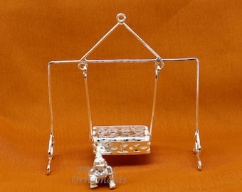 May include: A silver baby swing with a decorative design. The swing is hanging from a silver stand with a triangular top. A small silver figurine of a baby is sitting in the swing.