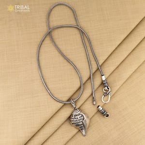 Op de afbeelding: Een zilveren ketting met een zilveren hanger in de vorm van een schelp. De schelp is gegraveerd met Sanskriet-tekst. De ketting heeft een karabijnhaak en een zilveren verlengketting.