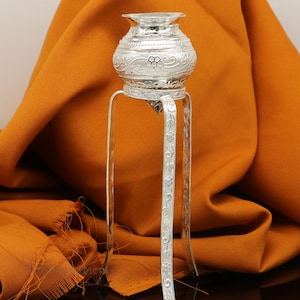 May include: A silver decorative pot with intricate floral designs sits on a three-legged stand. The stand is also silver and has a similar floral design.