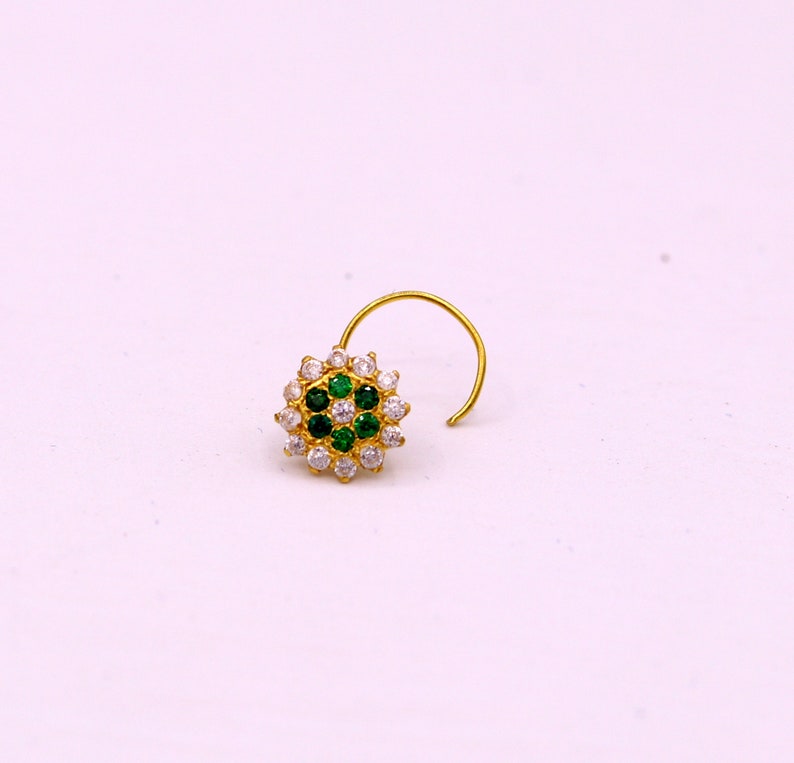 May include: A gold nose ring with a circular design featuring white and green gemstones.