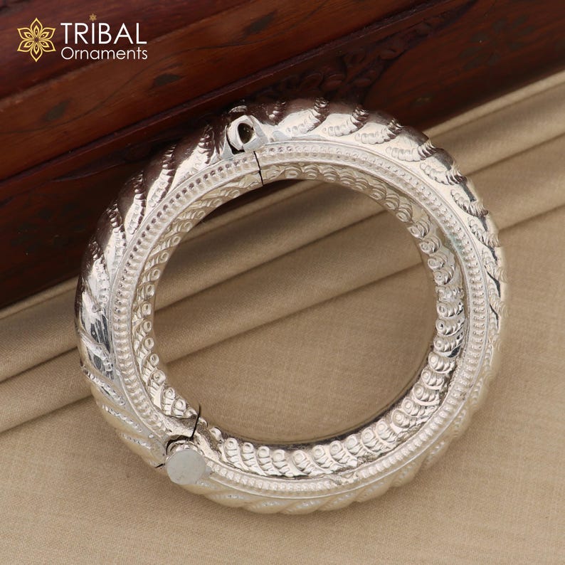 May include: A silver bangle bracelet with an intricate, textured design. The bracelet is open and has a hinged clasp.