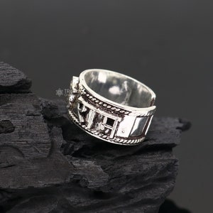 Peut inclure: Bague en argent avec des détails complexes et un motif en relief. La bague est placée sur un fond noir.