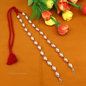 May include: A silver necklace with a red cord and tassel. The necklace is made of silver beads in a diamond shape. The necklace is on a brown surface.