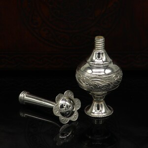 May include: A silver perfume bottle with an ornate design and a separate nozzle. The bottle has a round body and a short, wide neck. The nozzle is a long, thin tube with a flower-shaped end.