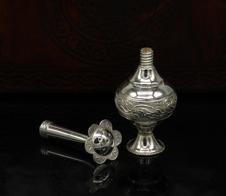 May include: A silver perfume bottle with an ornate design and a separate nozzle. The bottle has a round body and a short, wide neck. The nozzle is a long, thin tube with a flower-shaped end.