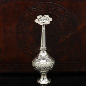 May include: A silver decorative bottle with a floral design and a long spout. The bottle is sitting on a dark brown surface.