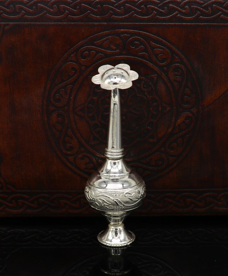 May include: A silver decorative bottle with a floral design and a long spout. The bottle is sitting on a dark brown surface.