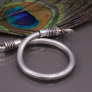 K&ouml;nnte beinhalten: Ein silbernes Schlangenkettenarmband mit strukturierter Oberfl&auml;che. Das Armband liegt auf einer Pfauenfeder.
