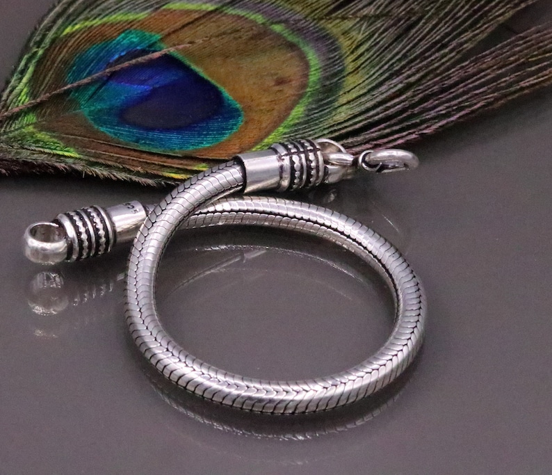 K&ouml;nnte beinhalten: Ein silbernes Schlangenkettenarmband mit strukturierter Oberfl&auml;che. Das Armband liegt auf einer Pfauenfeder.