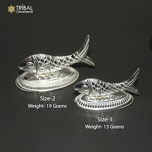 May include: Two silver fish ornaments. The larger fish, labelled "Size-2," weighs 19 grams. The smaller fish, labelled "Size-1," weighs 13 grams. Both fish sit on a decorative, circular base. The Tribal Ornaments logo is in the top left corner.