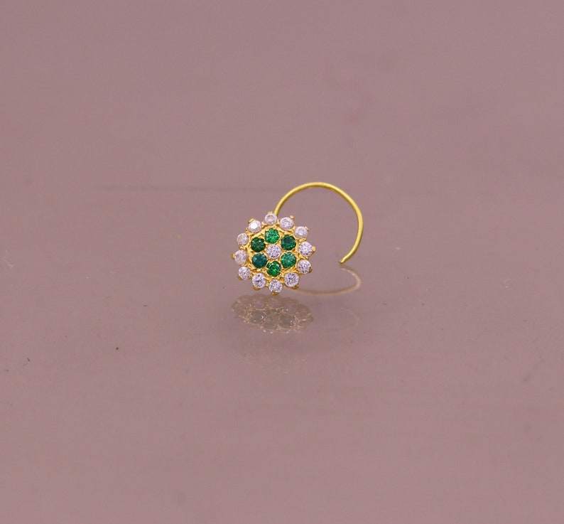 May include: A gold nose ring with a circular design featuring white and green gemstones.