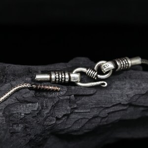 May include: Close-up of a silver chain necklace with a hook clasp. The chain is made of small, round links and has a textured finish. The clasp is marked with the number "925", indicating that it is made of sterling silver.