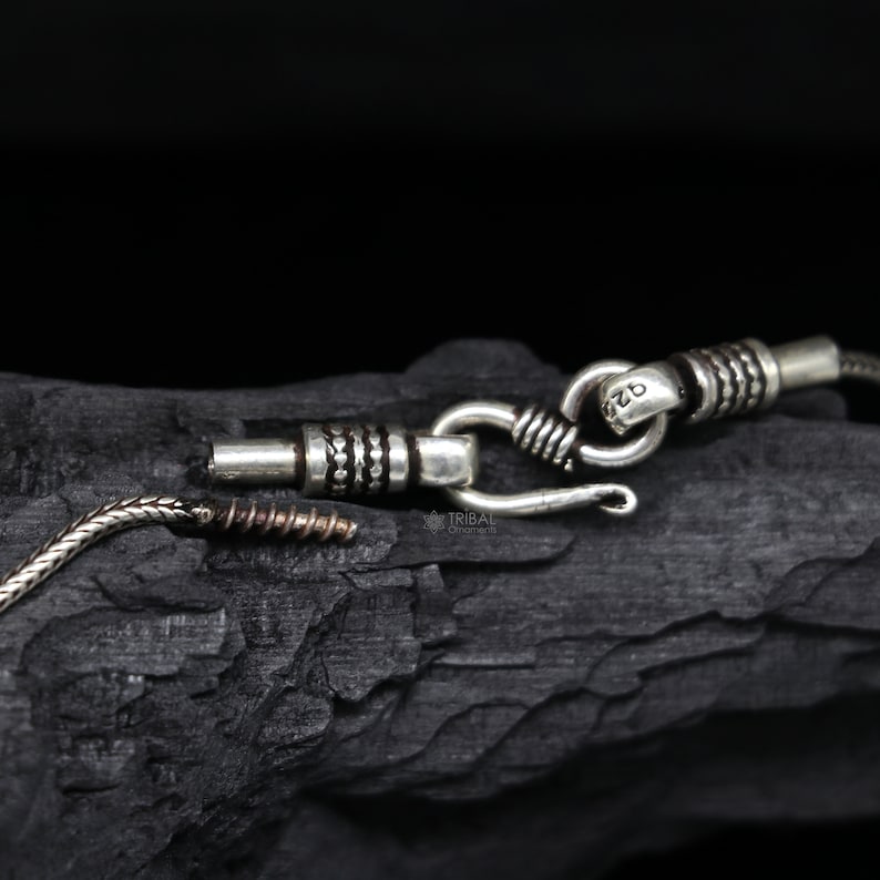 May include: Close-up of a silver chain necklace with a hook clasp. The chain is made of small, round links and has a textured finish. The clasp is marked with the number "925", indicating that it is made of sterling silver.